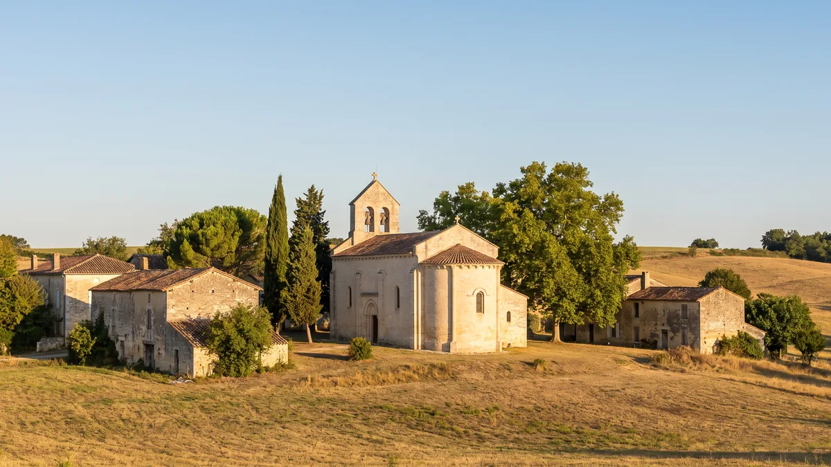 Una pequeña iglesia de pueblo de Lot-et-Garonne, construida en piedra clara, con campanario integrado en el muro, rodeada de casas de piedra y cipreses