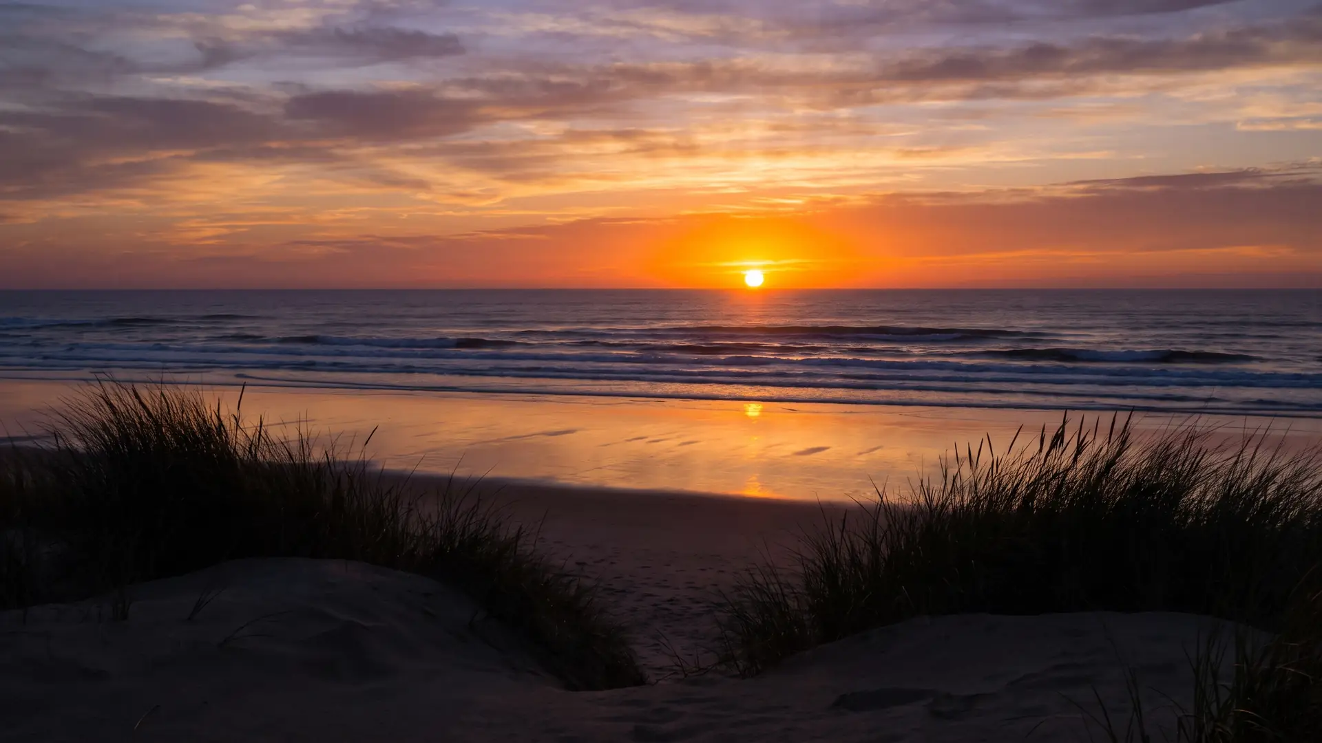 El sol poniente se funde en el Atlántico, la playa mojada refleja el resplandor, las dunas se perfilan en la silueta