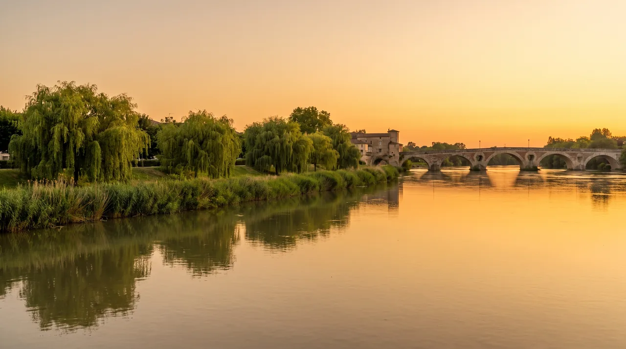 El río Garona, ancho y tranquilo, al atardecer cerca de Cadillac, con sus orillas arboladas y un puente metálico a lo lejos
