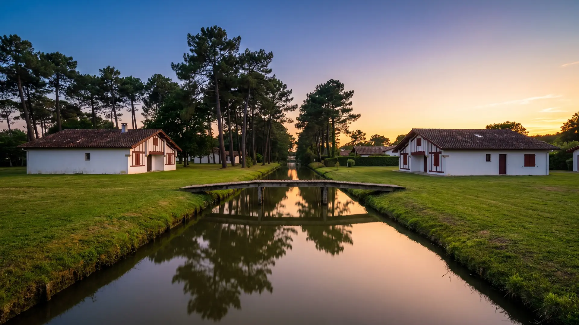 Un canal tranquilo bordeado de pinos y casas de las Landas con entramado de madera, una pasarela de madera a la hora dorada