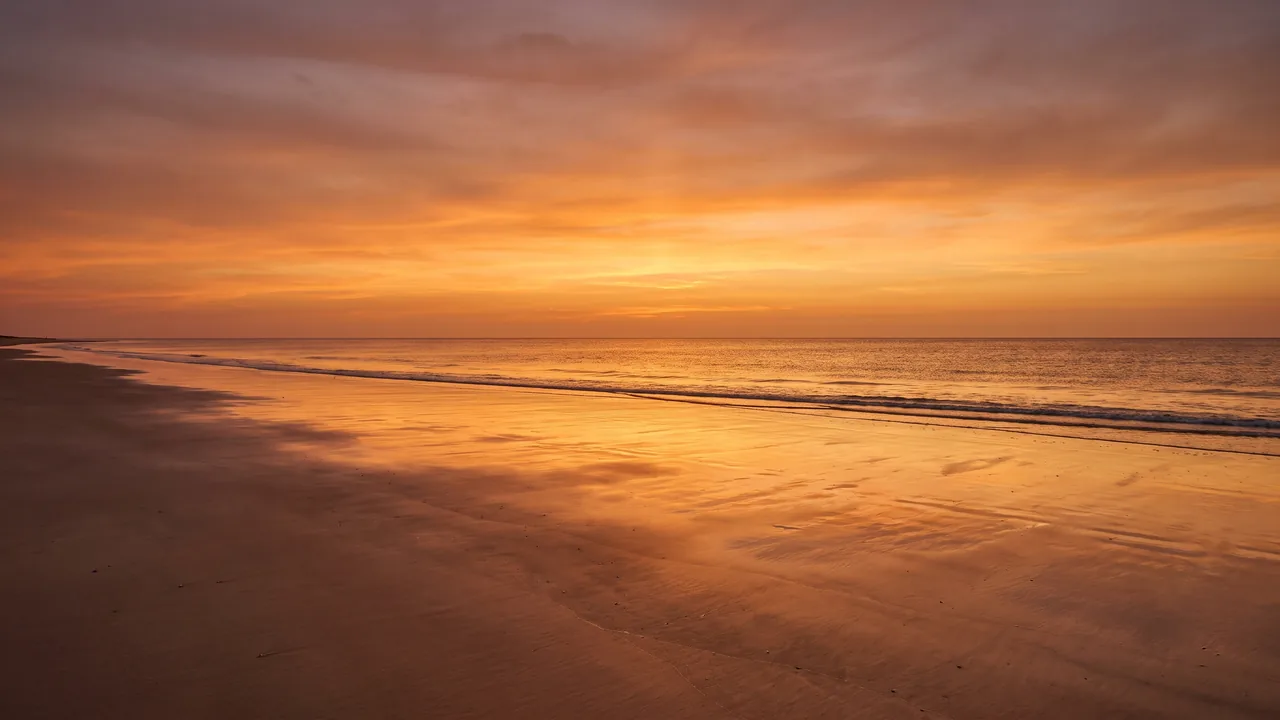 Puesta de sol anaranjada sobre el Atlántico desde una playa desierta de Carcans, con la arena húmeda reflejando el cielo cálido