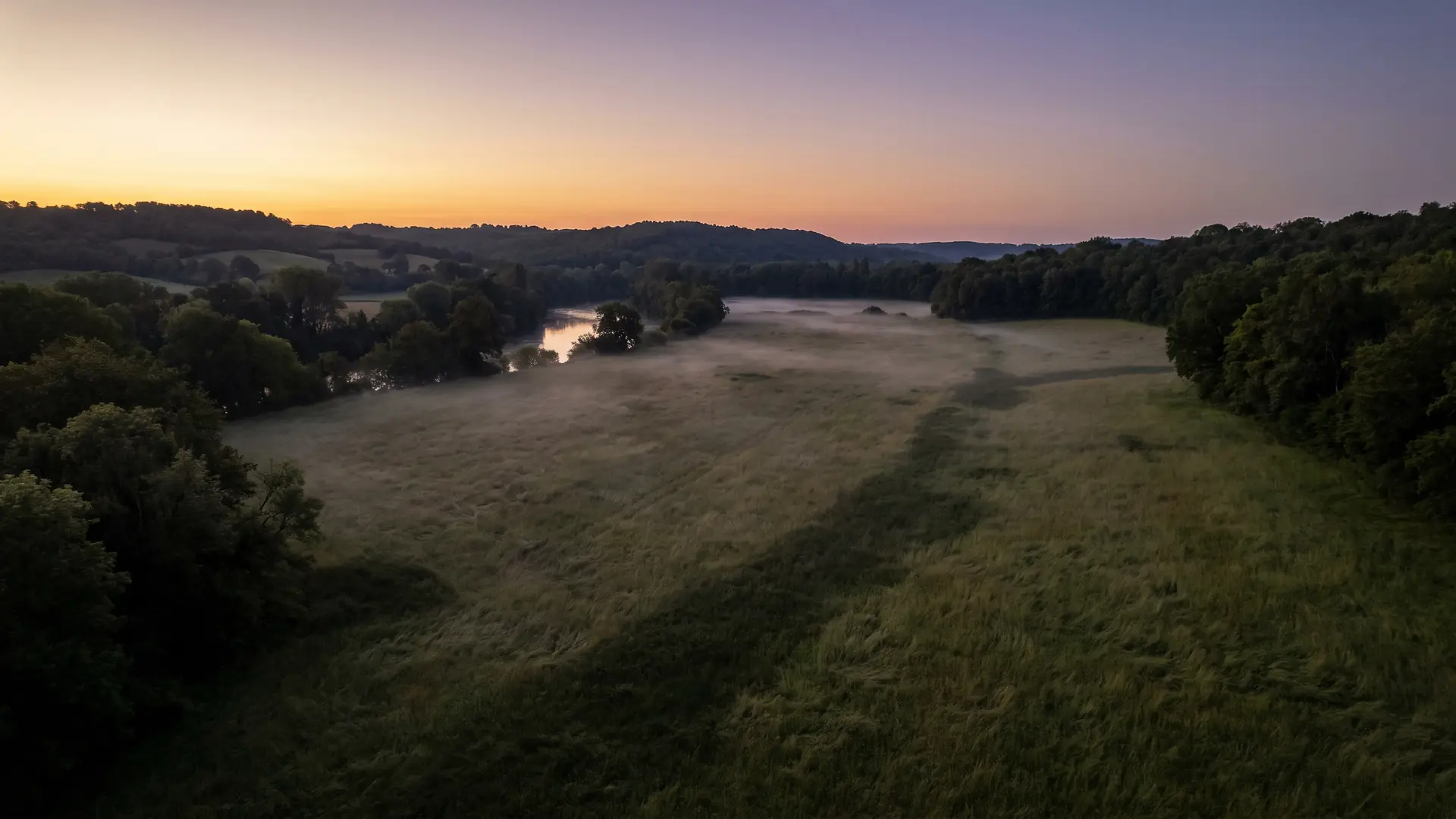 Una extensa pradera a orillas del río Dordoña al atardecer, con una ligera bruma y bosques oscuros en el horizonte: un paisaje solemne