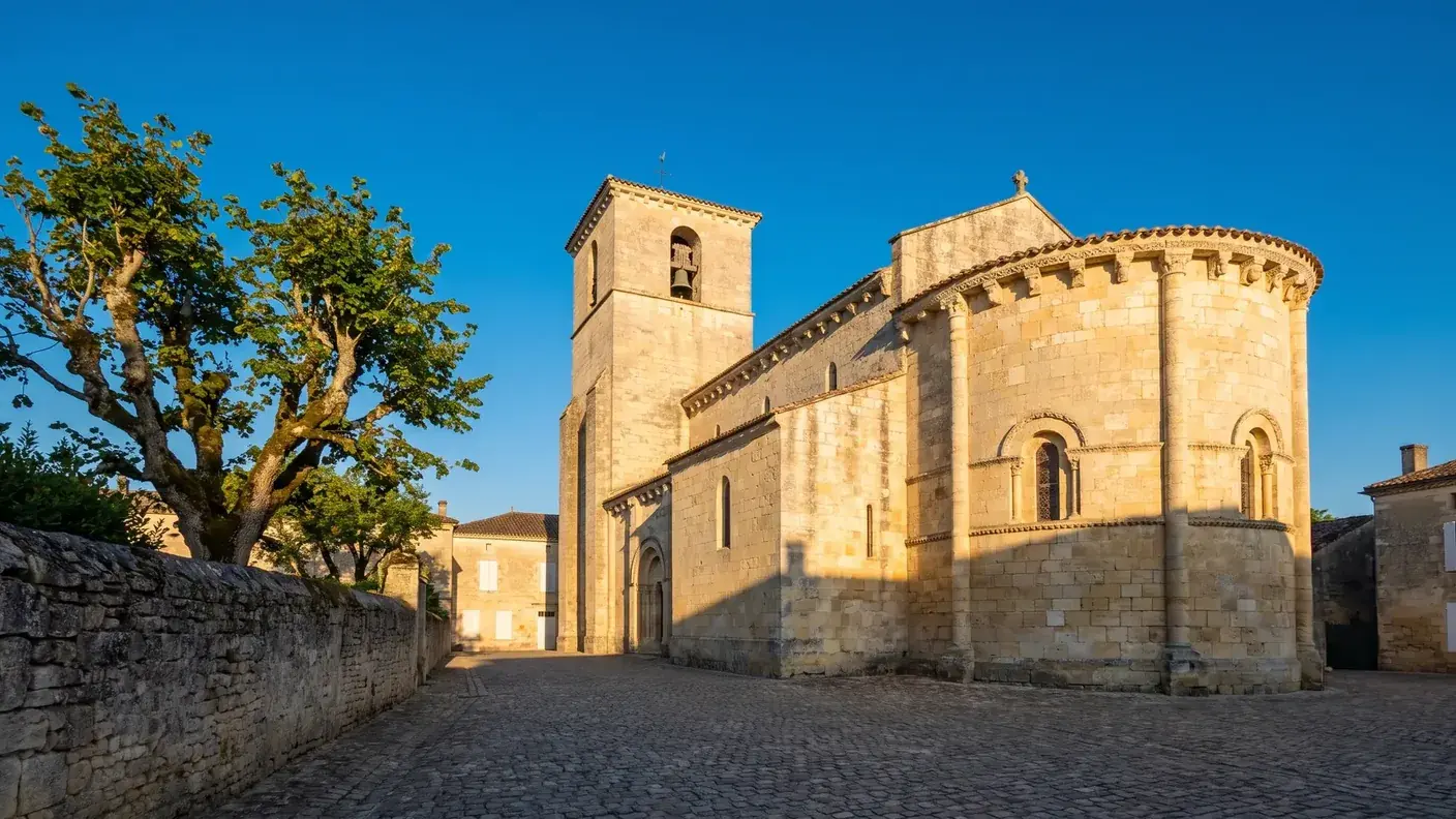 Una iglesia de pueblo construida en piedra caliza clara en Gironda, con cabecera redondeada, campanario cuadrado y una plaza rodeada de un tilo