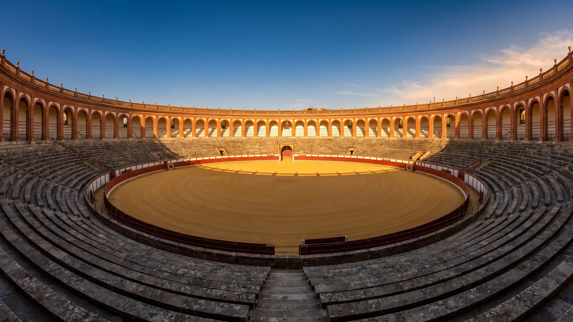 Unas arenas históricas vacías, de ladrillo y arcadas blancas, arena dorada y gradas desiertas a la hora dorada