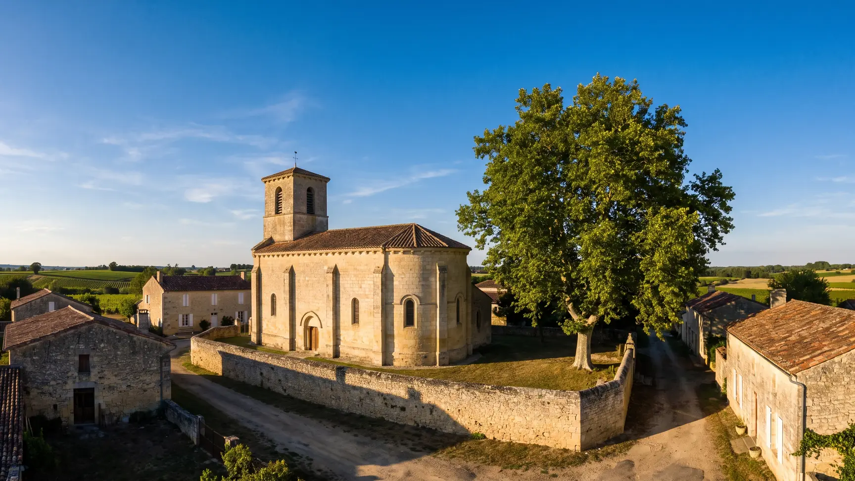 Una pequeña iglesia de pueblo, construida en piedra clara y con un campanario cuadrado, rodeada de casas bajas con tejados de tejas