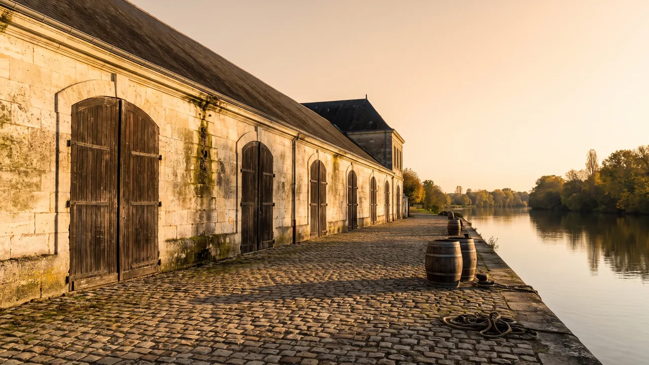 Larga fachada de piedra clara de una bodega de envejecimiento de coñac en Jarnac, con altas puertas arqueadas y tejado de tejas