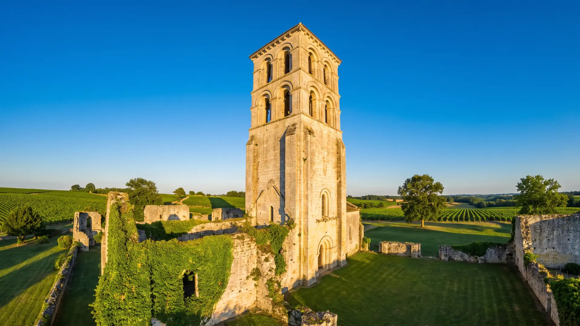 Un alto campanario de una abadía románica, construido en piedra clara, que se recorta contra el cielo azul sobre unas murallas en ruinas
