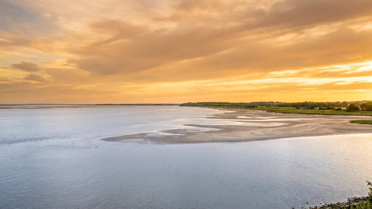 El vasto estuario de la Gironda al atardecer, una inmensa extensión de agua tranquila y plateada que se extiende hasta una orilla lejana