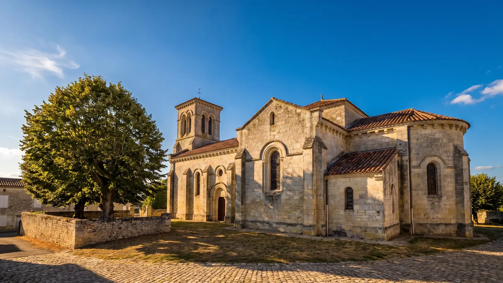 Una iglesia de pueblo del siglo XIX, construida en piedra clara, con torre-campanario y ventanas altas, situada en una plaza rodeada de un tilo