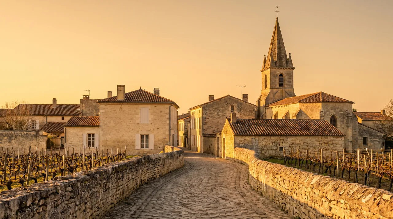 Una callejuela tranquila de un pueblo vinícola del Médoc, casas de piedra clara con contraventanas, un campanario al fondo, una luz cálida