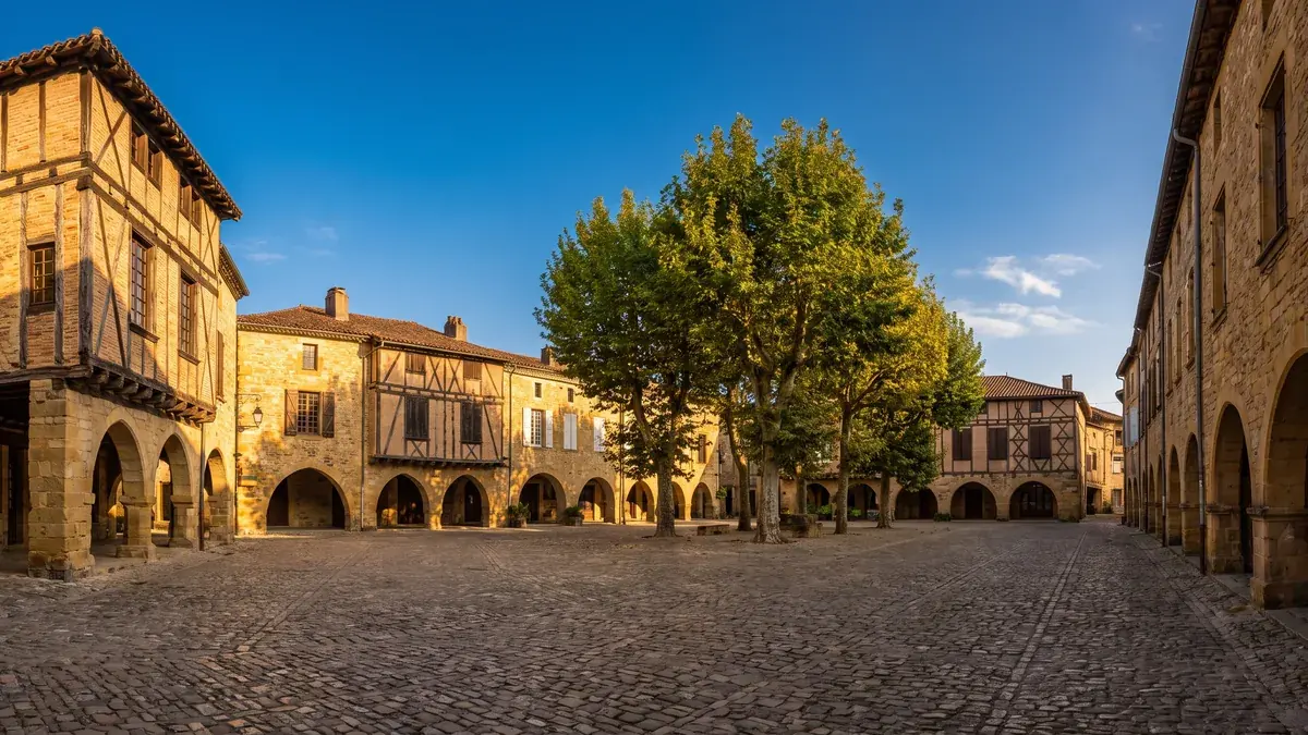 Una antigua plaza de Marmande rodeada de casas de piedra y con entramado de madera, con arcadas, tejados de tejas y plátanos.