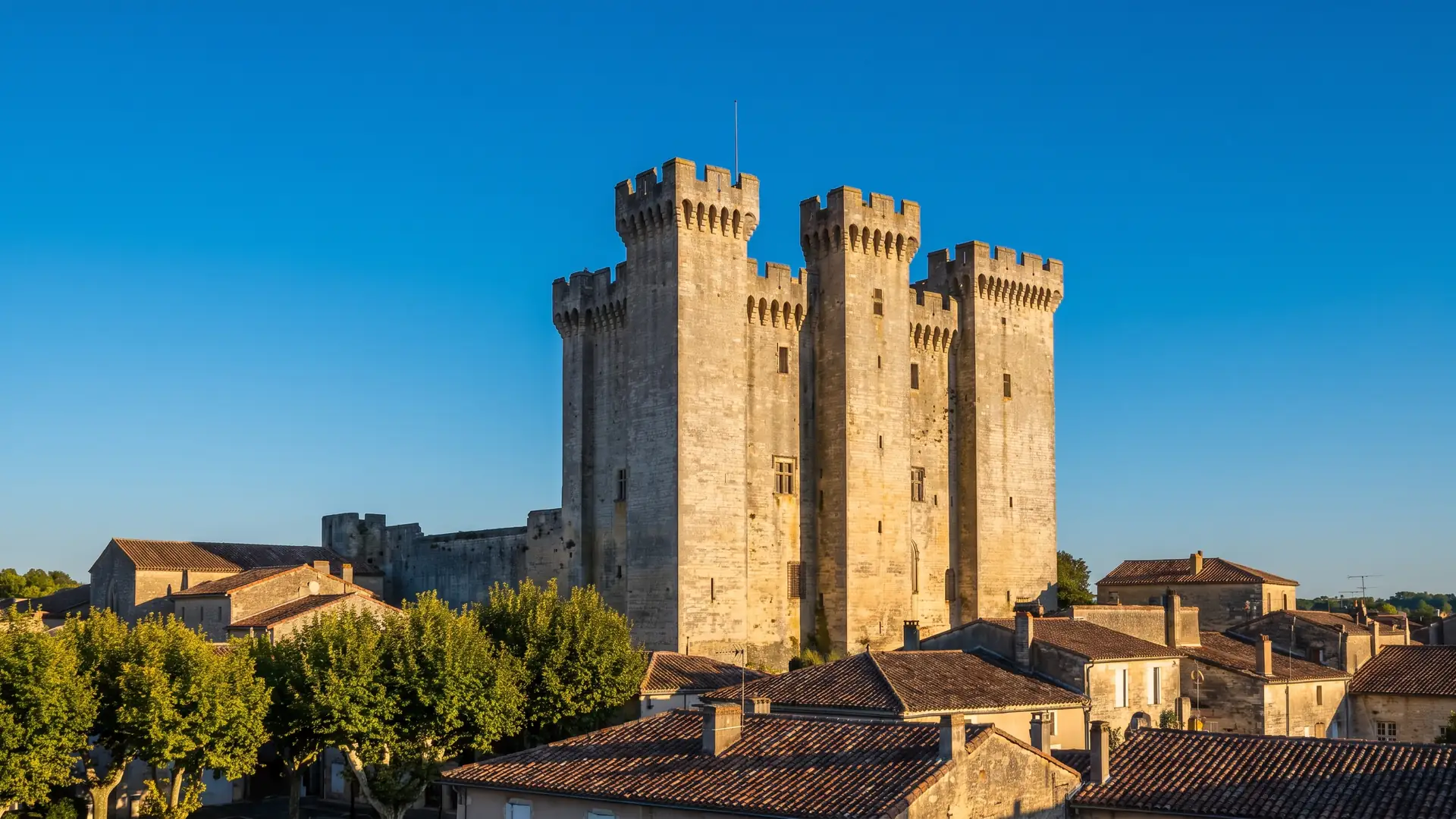 Una torre del homenaje medieval de planta cuadrada, con dos torres gemelas almenadas de piedra gris dorada, que se eleva sobre los tejados de una ciudad gascona