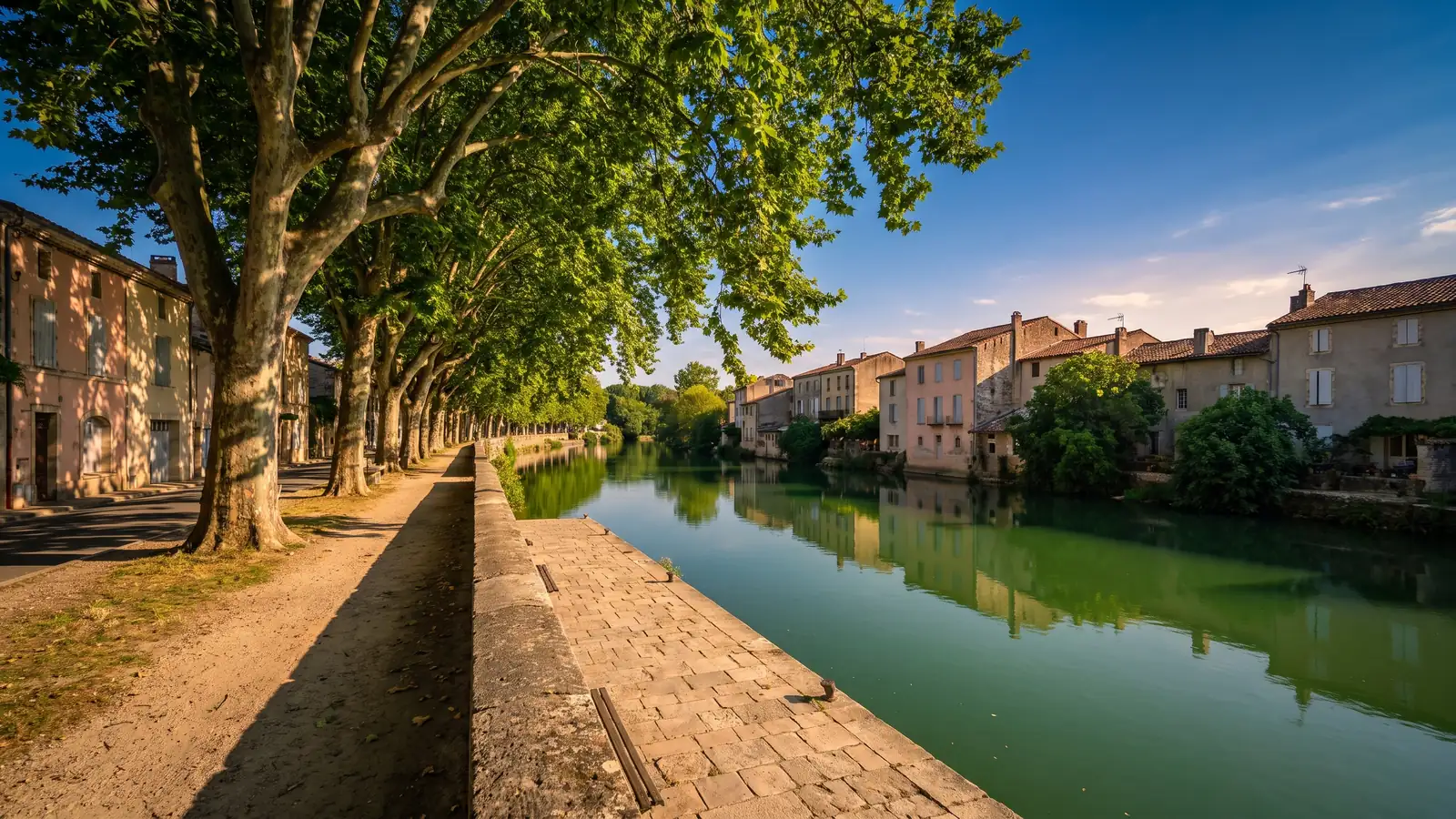 Un paseo a la sombra de los plátanos a orillas de un río verde, con muelles de piedra y casas de colores pastel reflejadas en el agua