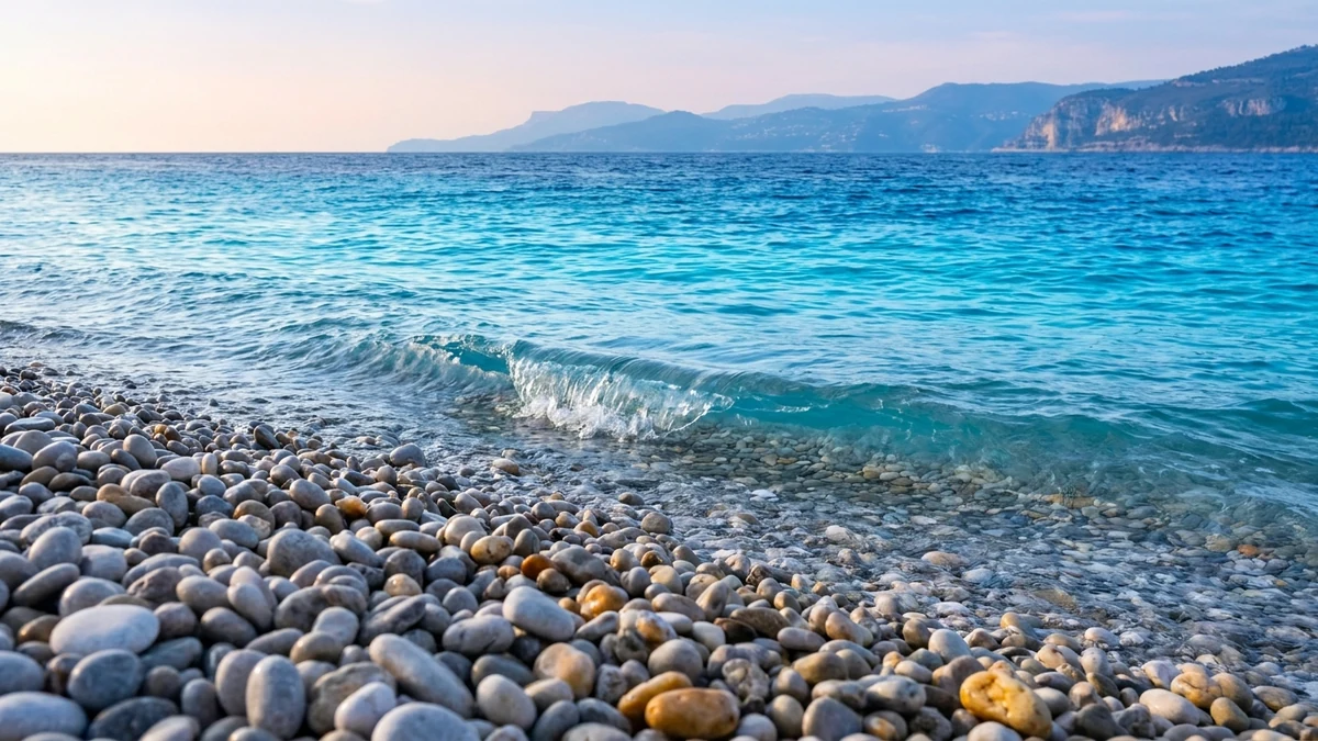 Plage de galets ronds gris et ambrés de la baie des Anges à Nice, vague transparente qui les recouvre, mer virant du turquoise au bleu profond vers l'horizon, collines bleutées au loin