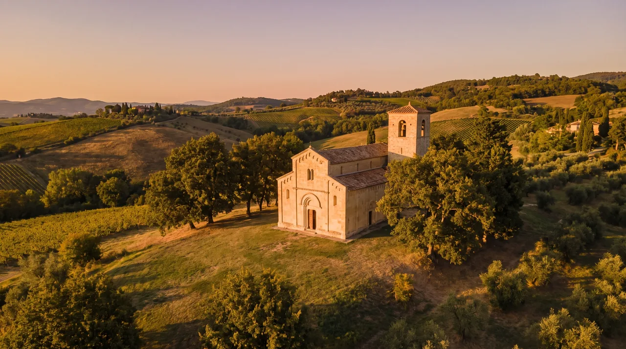 Iglesia románica de pueblo, construida en piedra clara, con campanario cuadrado y portal redondeado, árboles centenarios y la cálida luz del atardecer