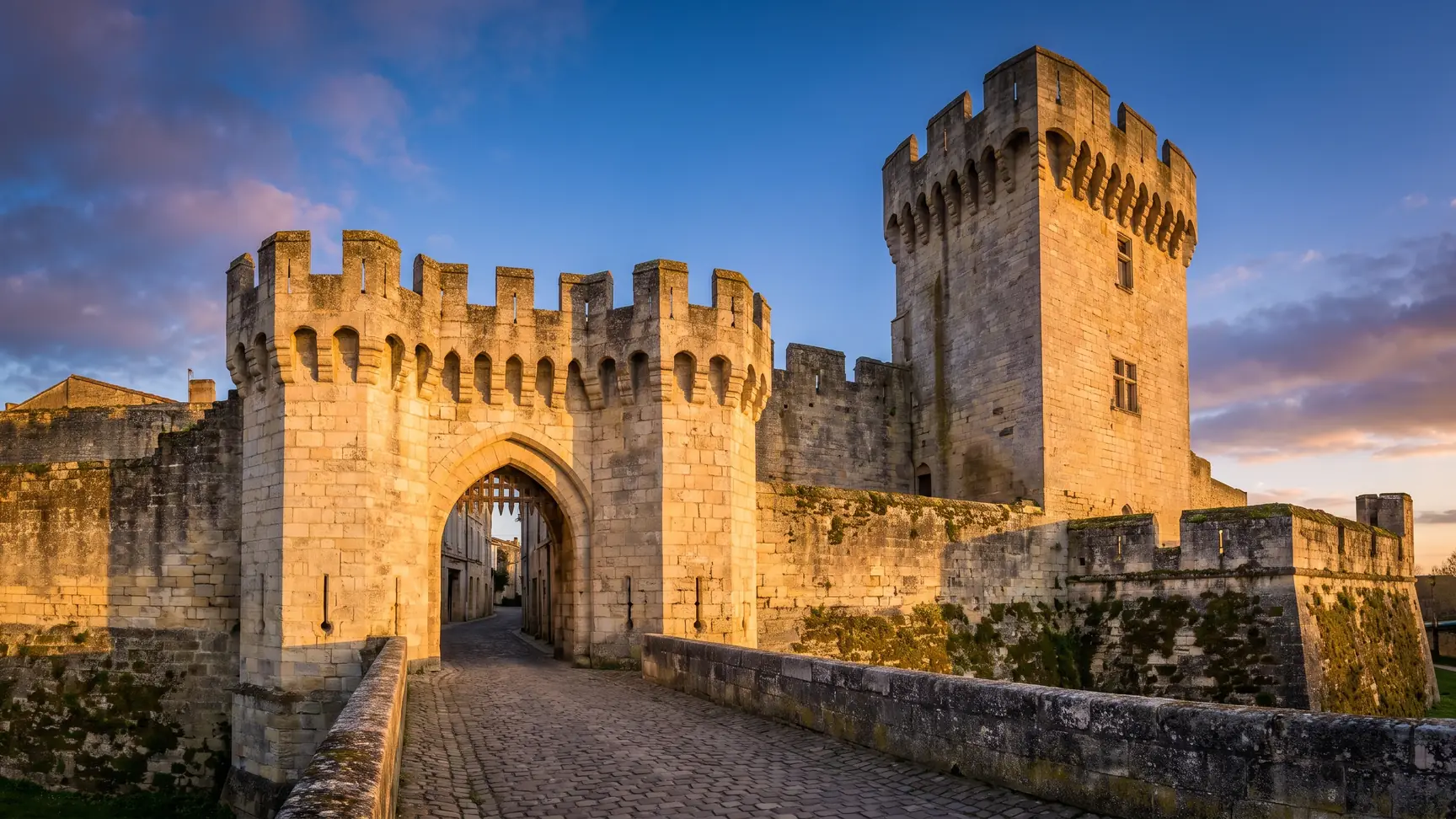 Una puerta fortificada medieval de piedra clara, flanqueada por una torre almenada, a la entrada de una ciudad amurallada