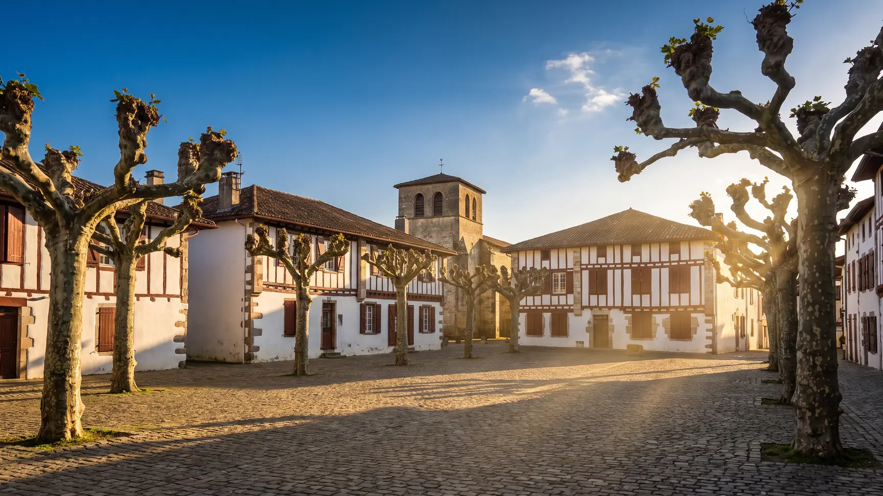 Una plaza de un pueblo de las Landas, a la sombra de plátanos podados, con casas bajas blancas con entramado de madera y el campanario de la iglesia al fondo